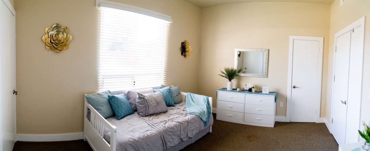 A cozy and well-lit bedroom featuring a daybed with pillows, a dresser with a mirror, and a decorative plant.