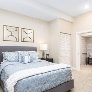 A modern bedroom with a neatly made bed, decorative pillows, and framed artwork above the headboard. A lamp on the nightstand provides soft lighting, and a doorway leads to an adjoining room.