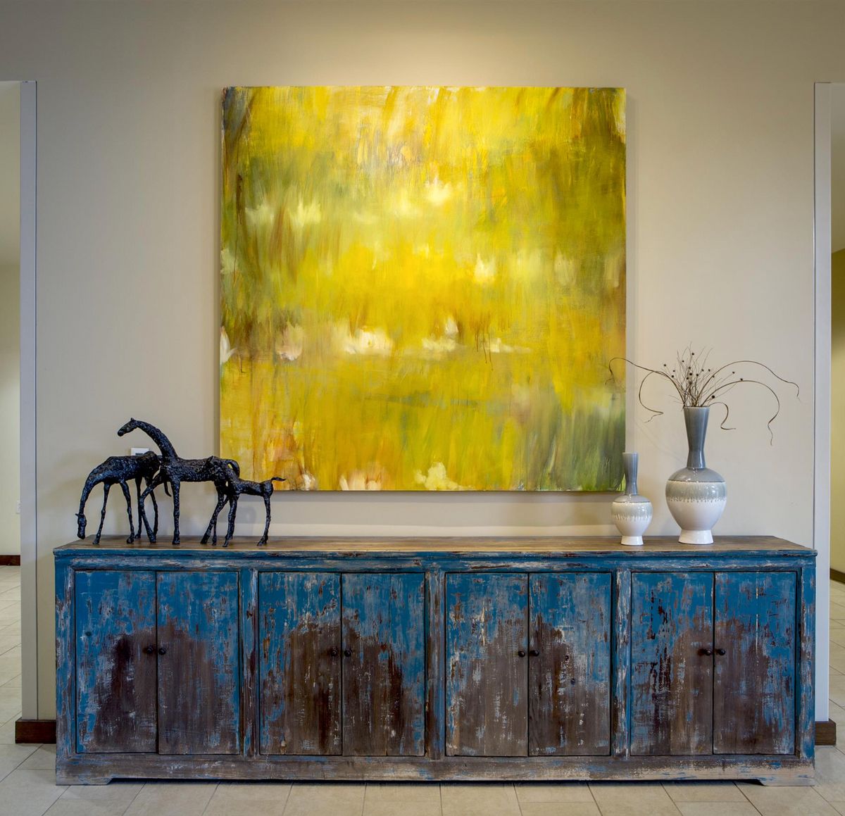A cozy interior scene featuring a textured blue console table adorned with decorative horse figurines and a vase, set against the backdrop of a vibrant yellow abstract painting.