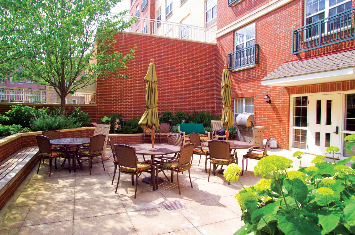 A cozy outdoor patio area with multiple tables and chairs, shaded by umbrellas, surrounded by a brick building and greenery.
