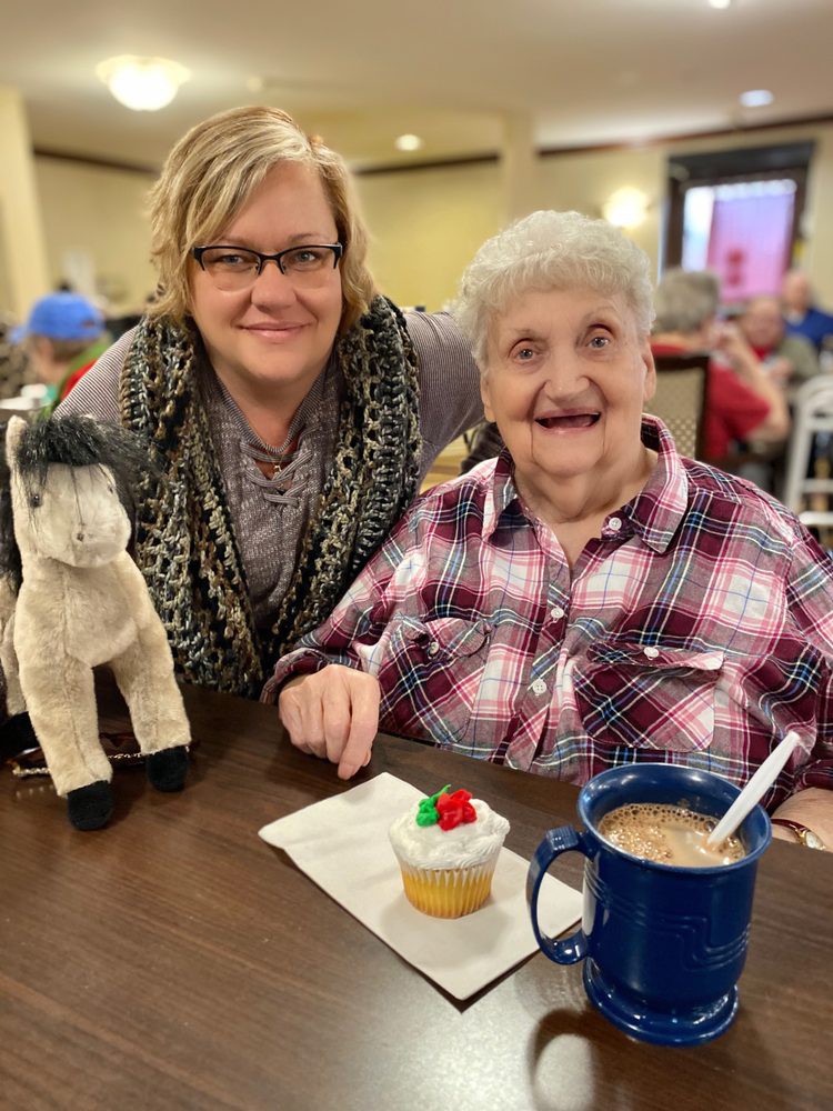 An elderly woman enjoying a cupcake and coffee, smiling alongside a companion indoors.