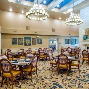 A spacious dining room with elegant chandeliers, round tables, and patterned carpet.