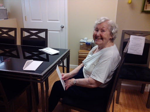 An elderly woman smiles while sitting at a table indoors.