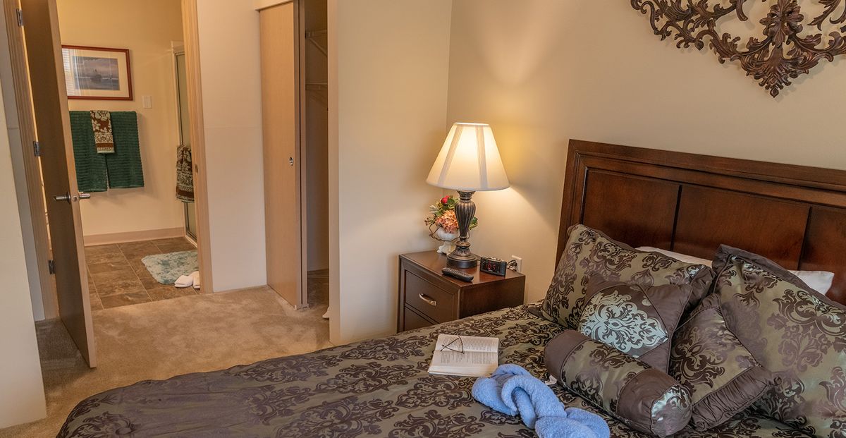 A cozy bedroom featuring a lamp on a nightstand beside a neatly made bed with patterned bedding and a book.