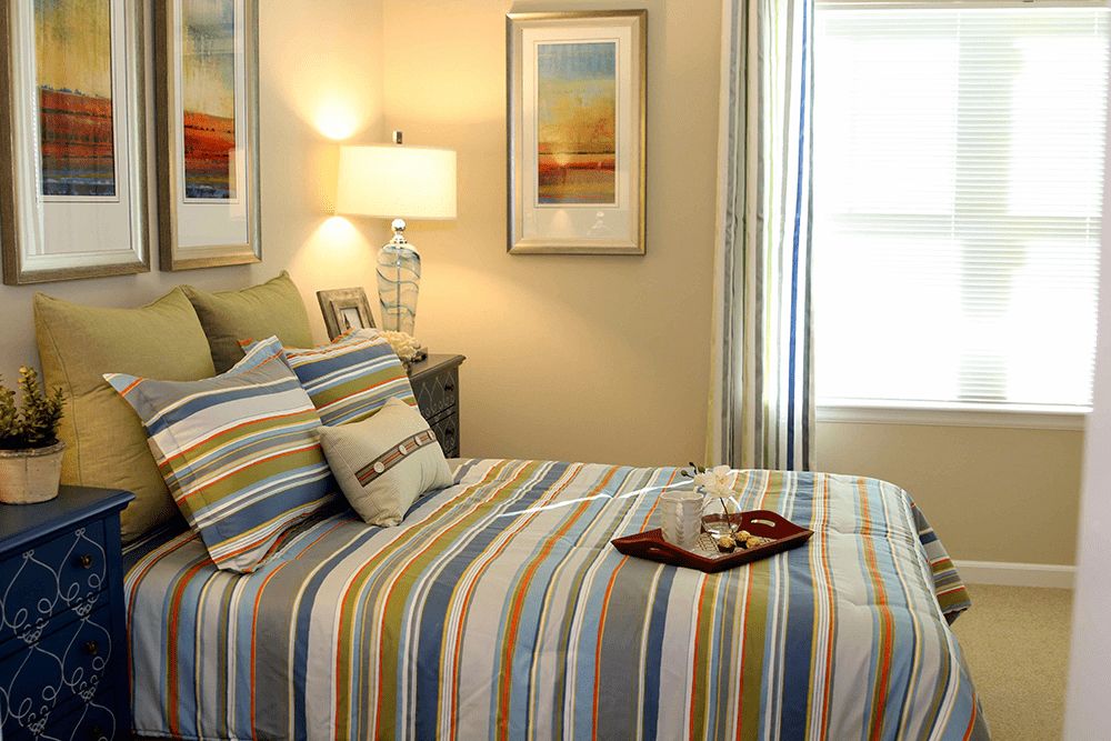 A cozy bedroom with striped bedding, decorative pillows, framed wall art, and a table lamp.