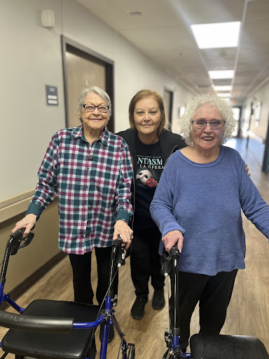 Two elderly women assisted by a caregiver as they walk down the hallway.