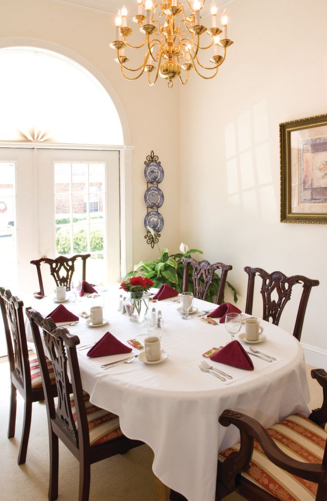 A bright dining room with a round table set for a meal, featuring red napkins and a centerpiece of flowers.