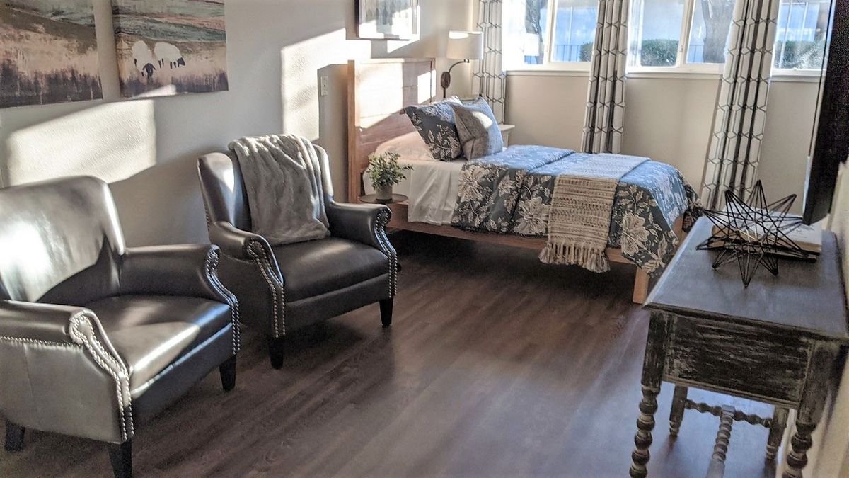 A cozy bedroom with two armchairs, a bed, and sunlight streaming through the window.
