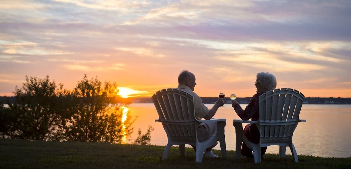 A couple enjoys a peaceful sunset by the lake, toasting with glasses while seated in outdoor chairs.