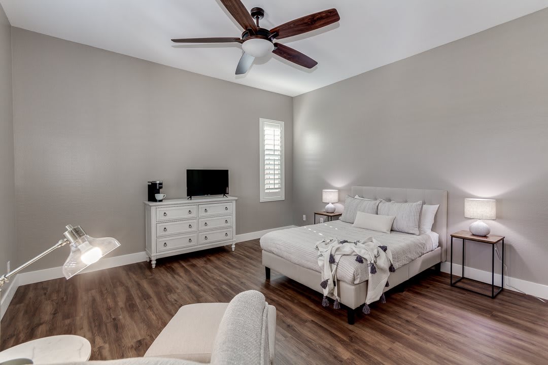 A modern bedroom interior featuring a neatly made bed, dresser with a TV, and a ceiling fan.