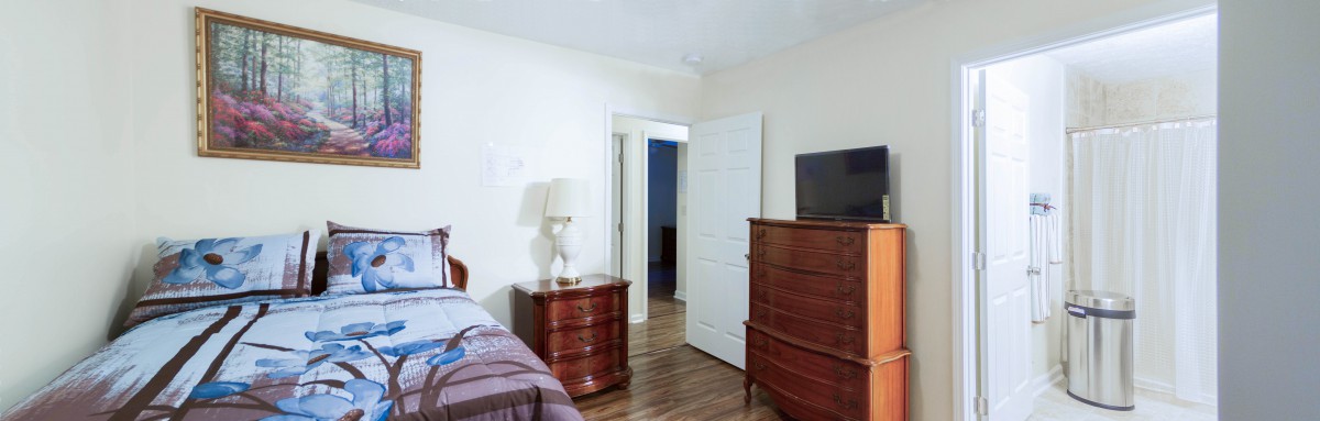 A bedroom featuring a dresser with a television and a bedside table with a lamp.