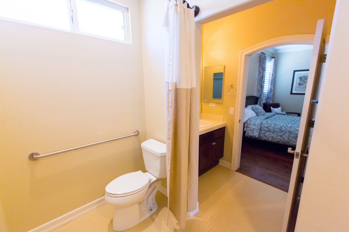 A bathroom with a shower, toilet, and sink, leading into a bedroom.