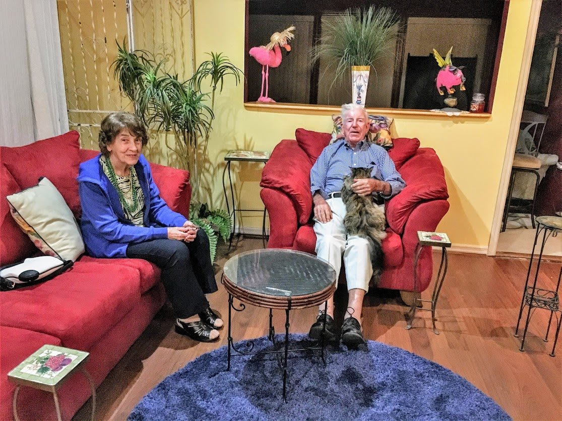 Two people sitting on red sofas in a cozy living room with plants and a dog.