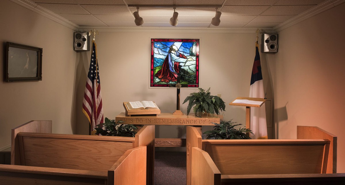 A chapel interior featuring a pulpit with an open Bible, stained glass window, and flags.
