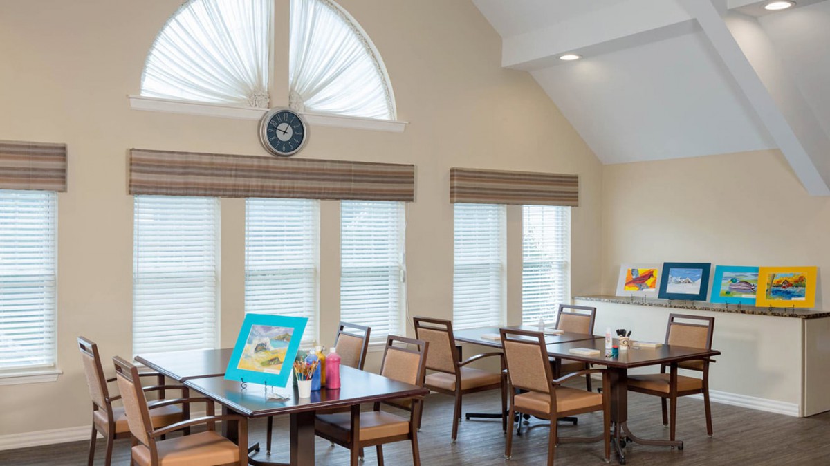 A well-lit room with several tables and chairs, featuring large windows and decorative elements including artwork and a clock.