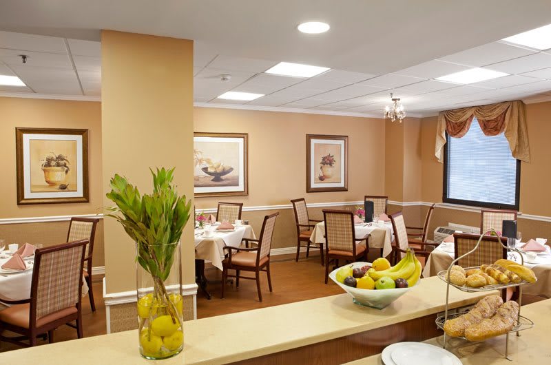 A cozy dining room with neatly arranged tables and chairs, decorated with art on the walls and a bowl of fresh fruit on a counter.