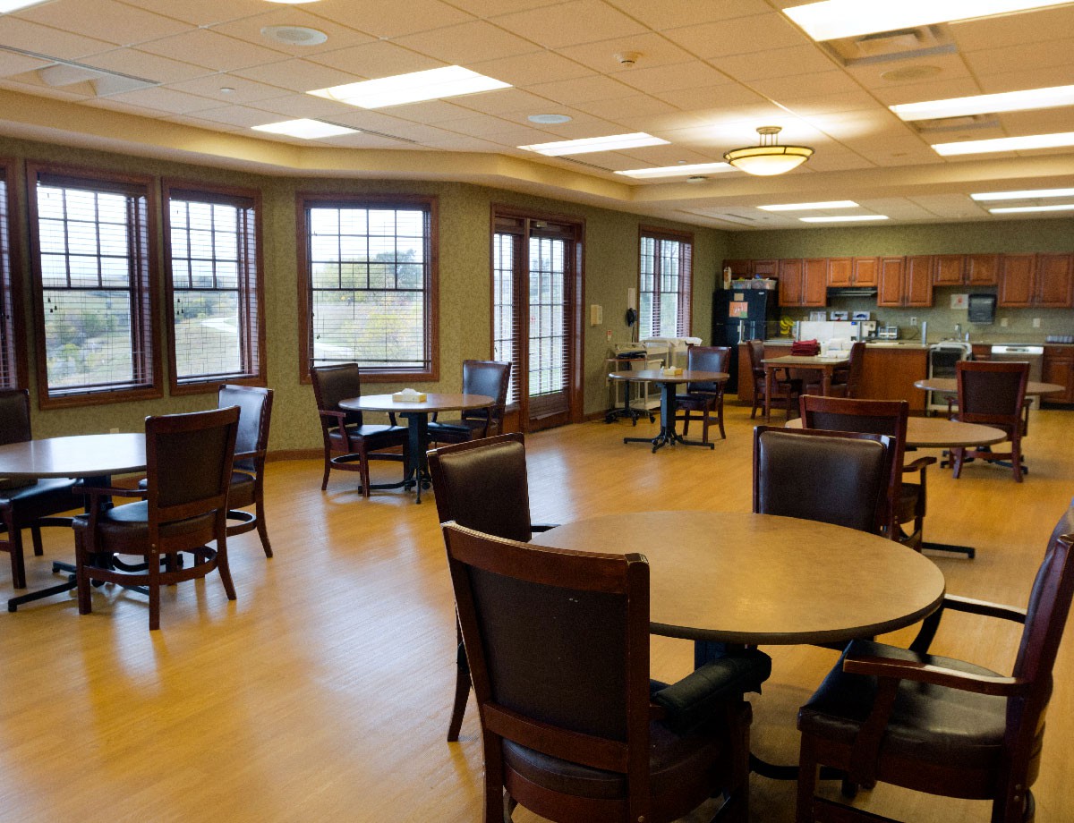 A spacious dining area with multiple tables and chairs, adjacent to a kitchen with large windows providing natural light.
