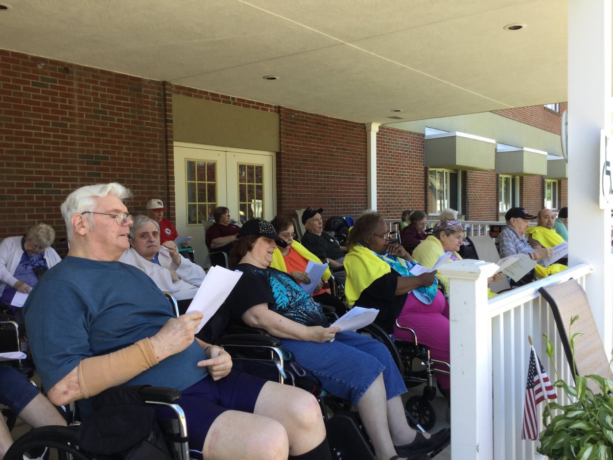 A group of people seated on a porch, some in wheelchairs, engaging in a reading activity.
