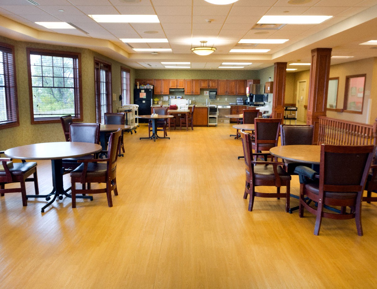 A spacious dining area with wooden flooring, multiple tables and chairs, and a kitchen in the background.