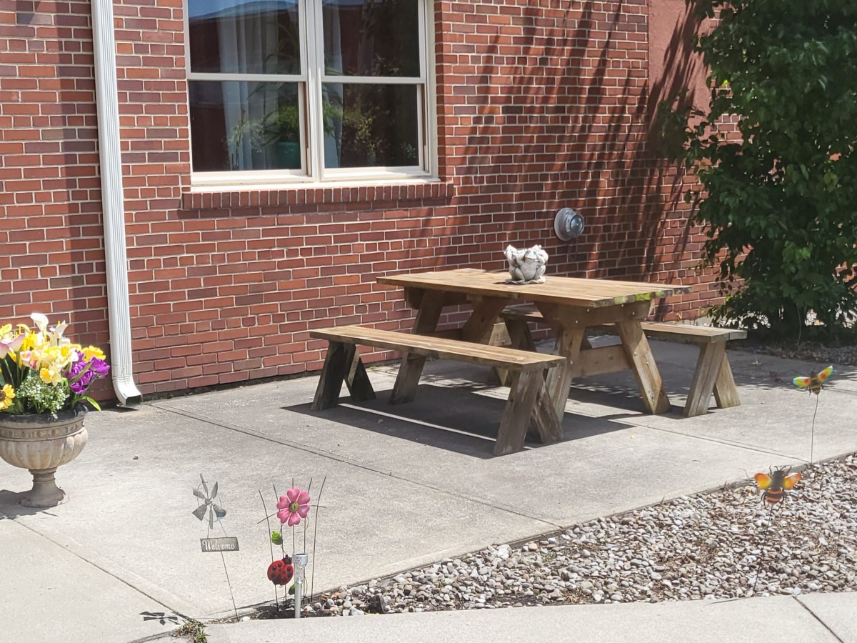 A wooden picnic table sits on a sunny patio next to a brick building.