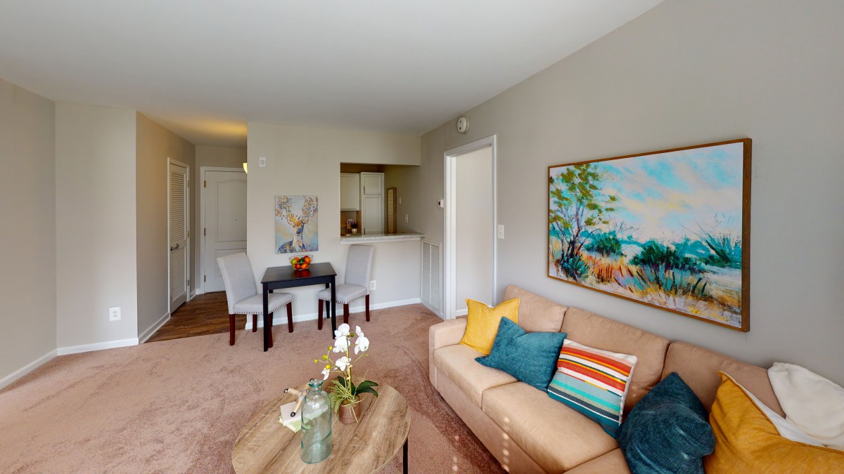 A cozy living room with a sofa, colorful pillows, artwork on the wall, and a dining table set.