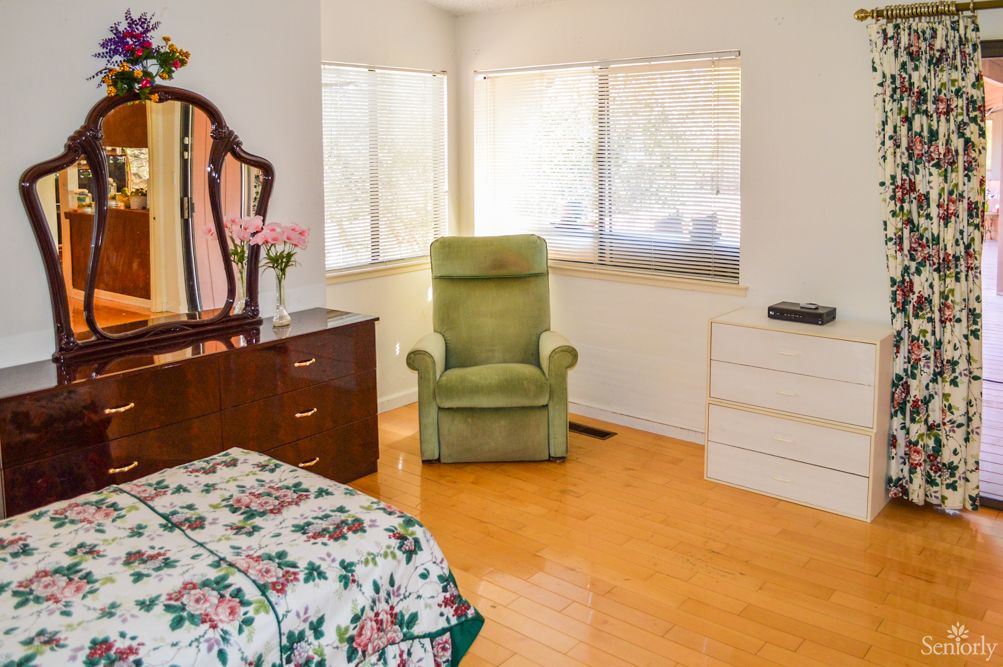 A cozy bedroom with vintage decor featuring a green armchair, floral bedspread, and natural light streaming through the windows.