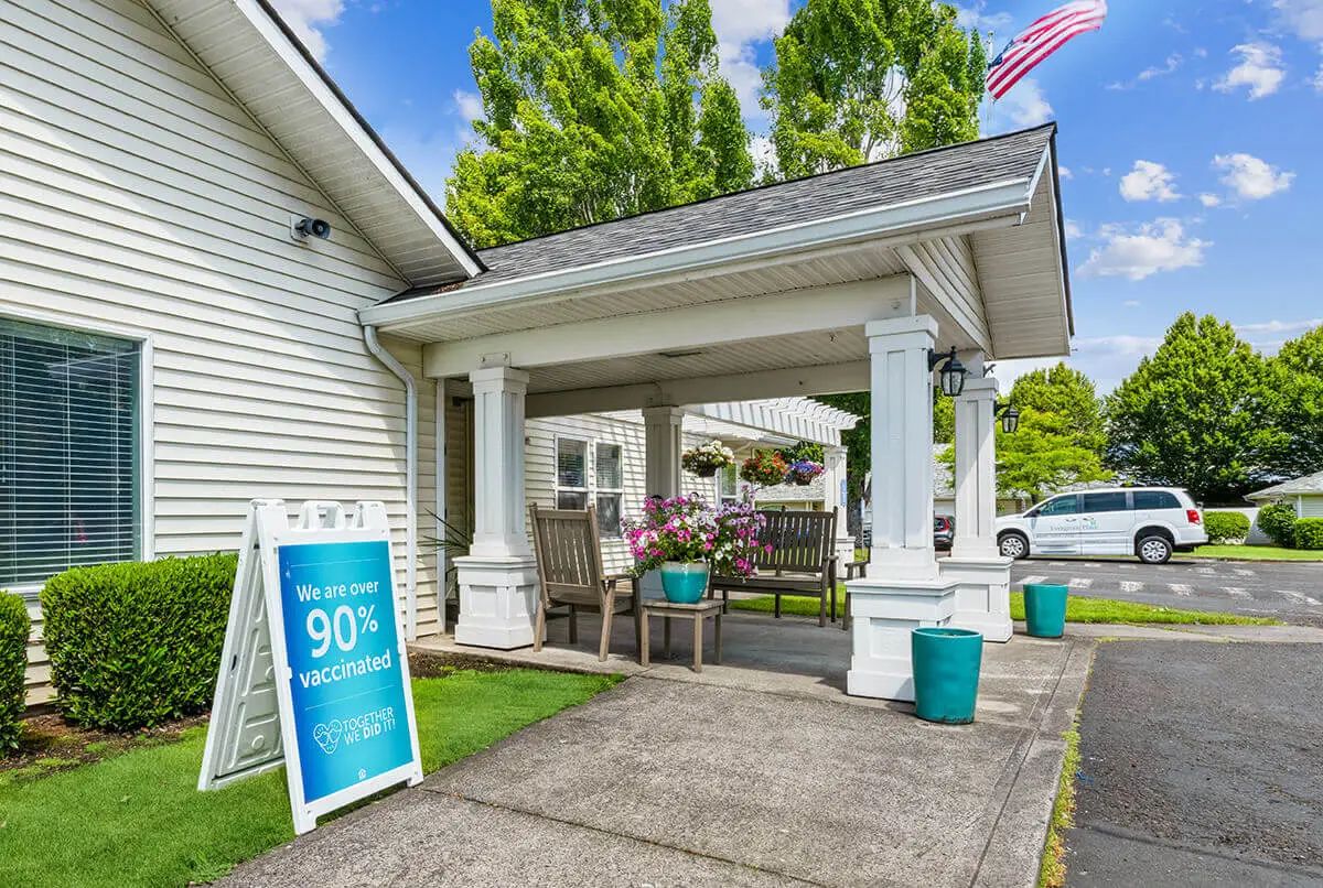 A welcoming entrance with a porch featuring outdoor seating and a signboard, flanked by greenery and an American flag.