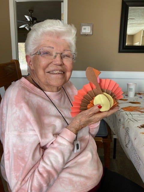 An elderly woman is smiling while holding a Thanksgiving-themed craft indoors.