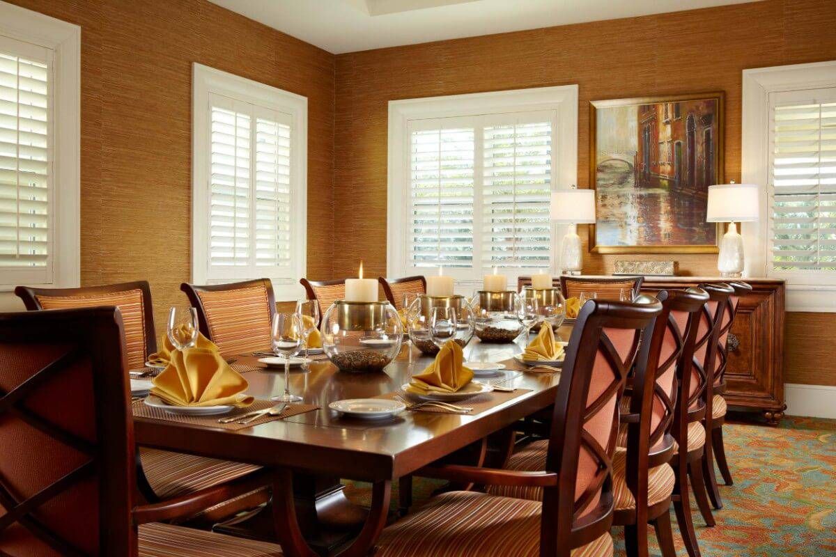 A beautifully set dining table in an elegant dining room with warm lighting and window shutters.