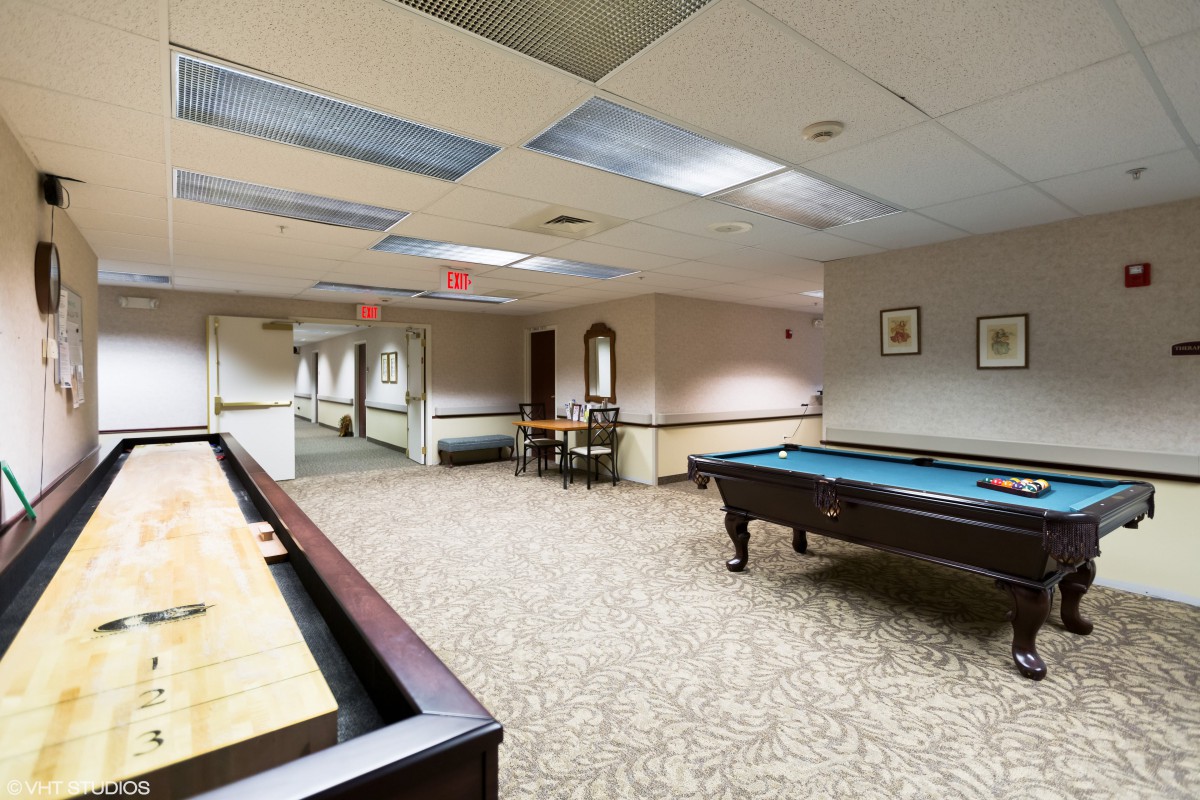 A well-lit game room featuring a pool table and shuffleboard, with carpeted flooring and ceiling lights.