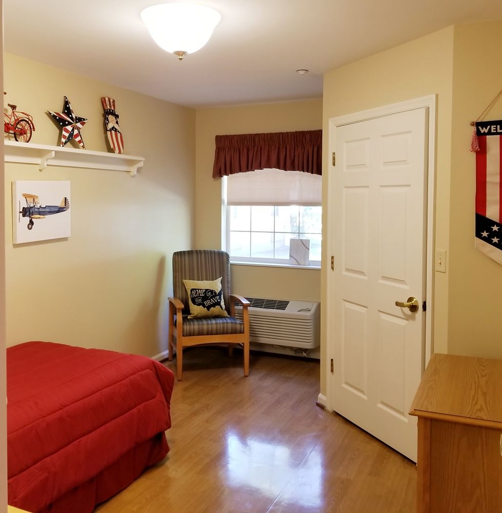 A small, cozy bedroom with wooden flooring, featuring a bed with red bedding, a wicker chair by the window, and wall decorations.