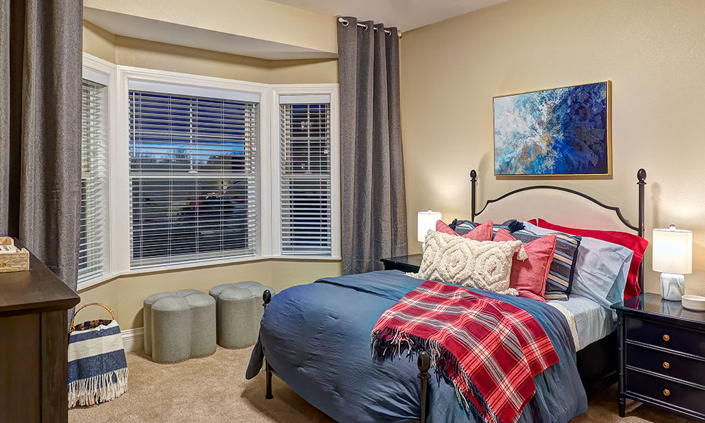 A cozy, well-decorated bedroom featuring a neatly made bed with colorful pillows and a plaid blanket, soft lighting, and a large window with blinds.