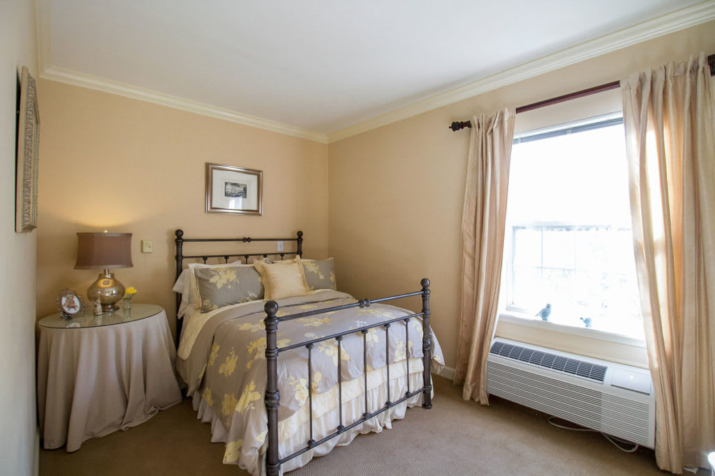 A cozy bedroom with a neatly made bed, bedside lamp, and sunlight streaming through the window.