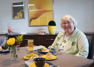 An elderly woman smiling while seated at a table with a meal in a yellow and green themed dining area.