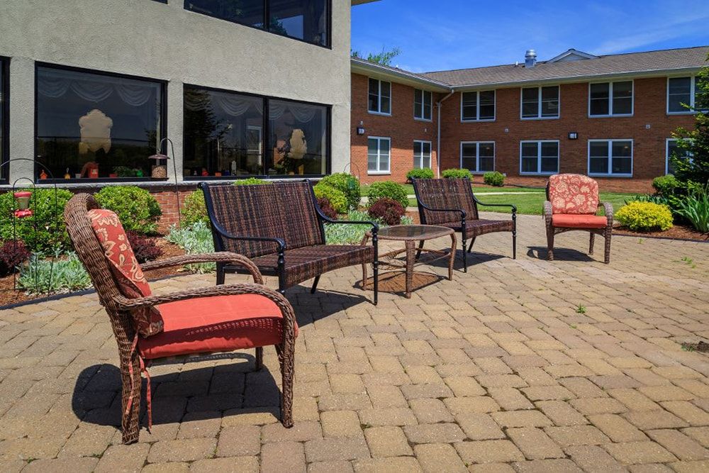 A sunny outdoor patio area with wicker chairs and tables on a brick surface, adjacent to a building.