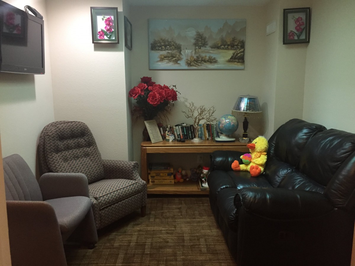 A cozy living room with a sofa and armchair, decorated with paintings, flowers, and a stuffed toy.