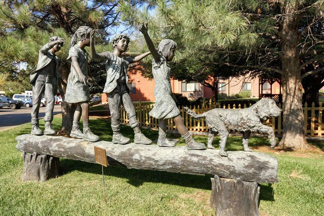 A bronze sculpture of children and a dog balancing on a log in a park setting.