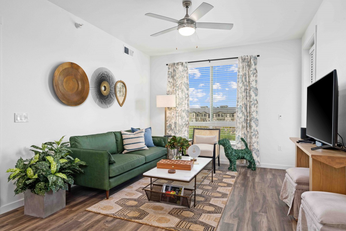 A modern living room with a green sofa, wooden flooring, and a large window letting in natural light. The space is decorated with indoor plants and wall art.