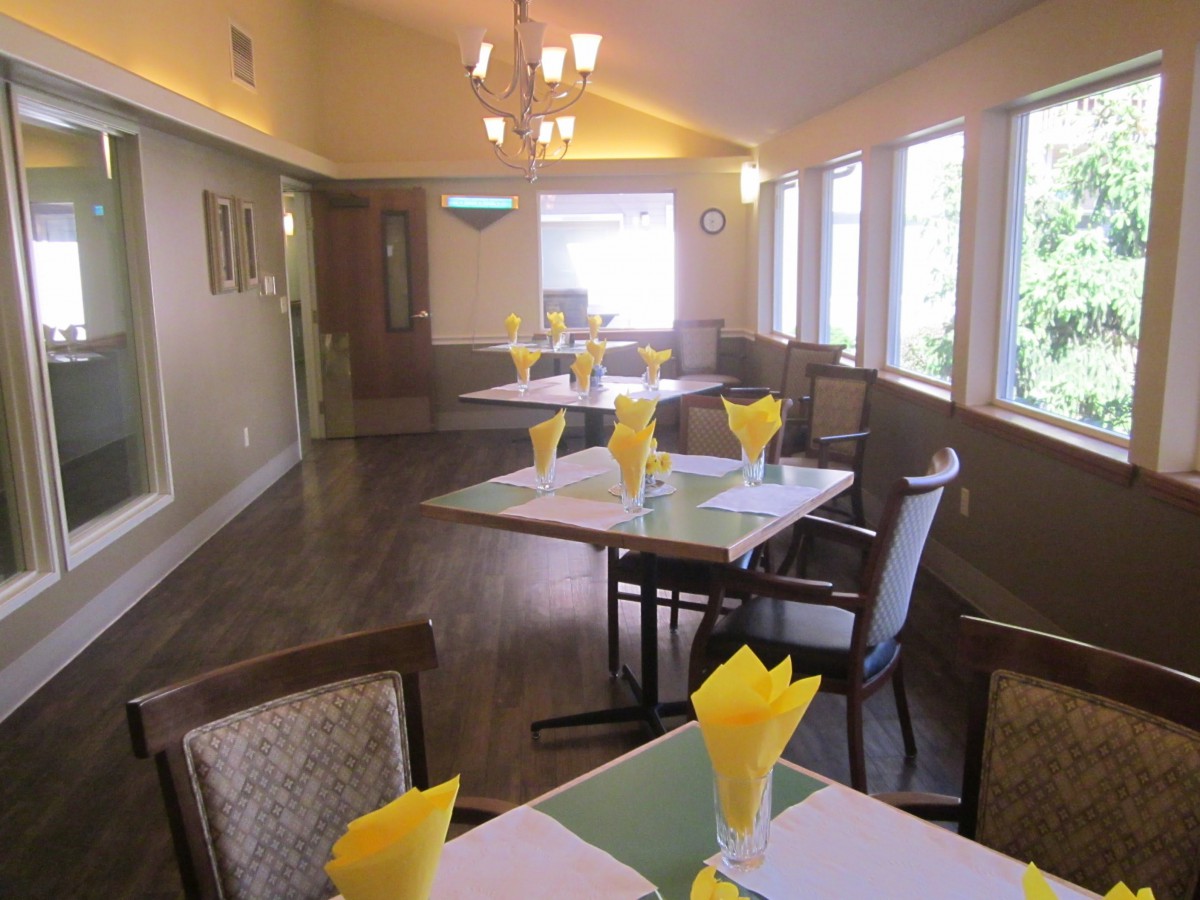 A cozy dining room with tables set for a meal, featuring yellow napkins and ample natural light from large windows.