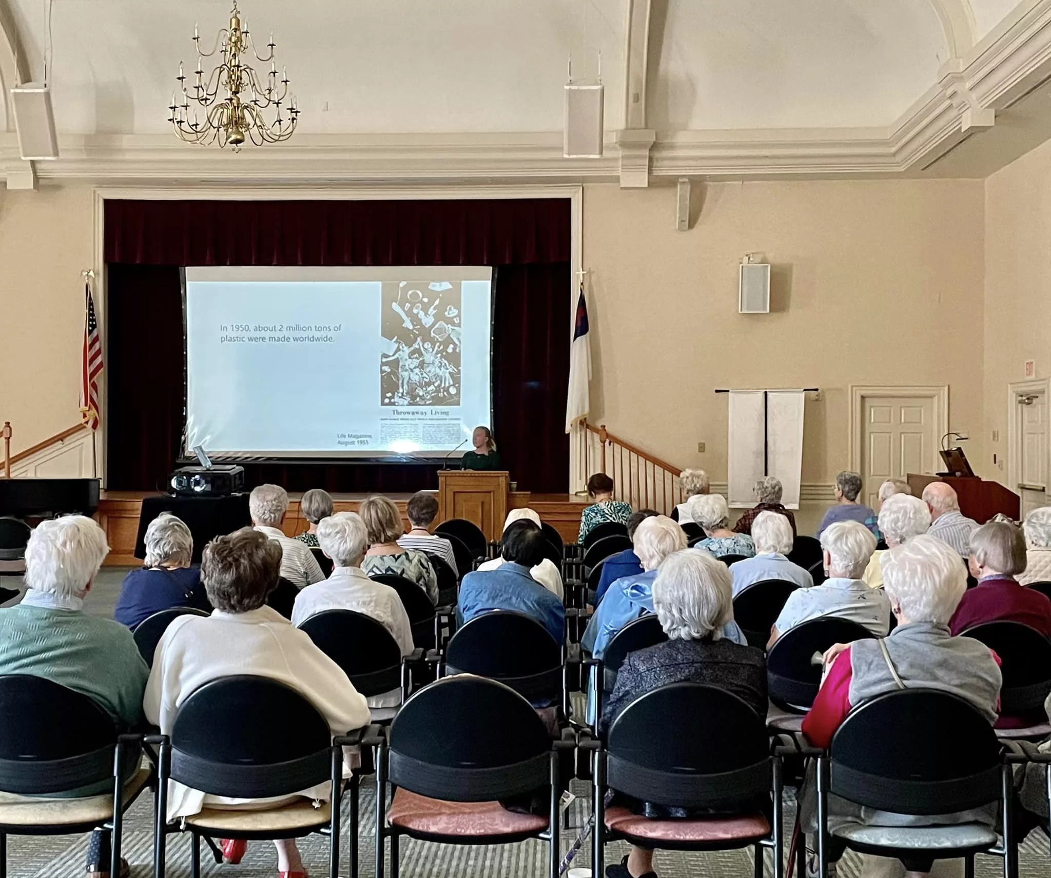 An audience seated in a hall watching a presentation on a large screen.