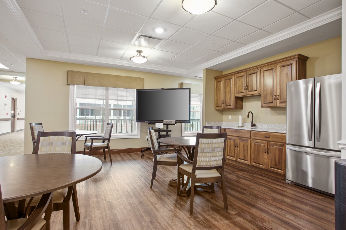 A cozy dining area with a kitchenette and television, featuring wooden furniture and a hardwood floor.