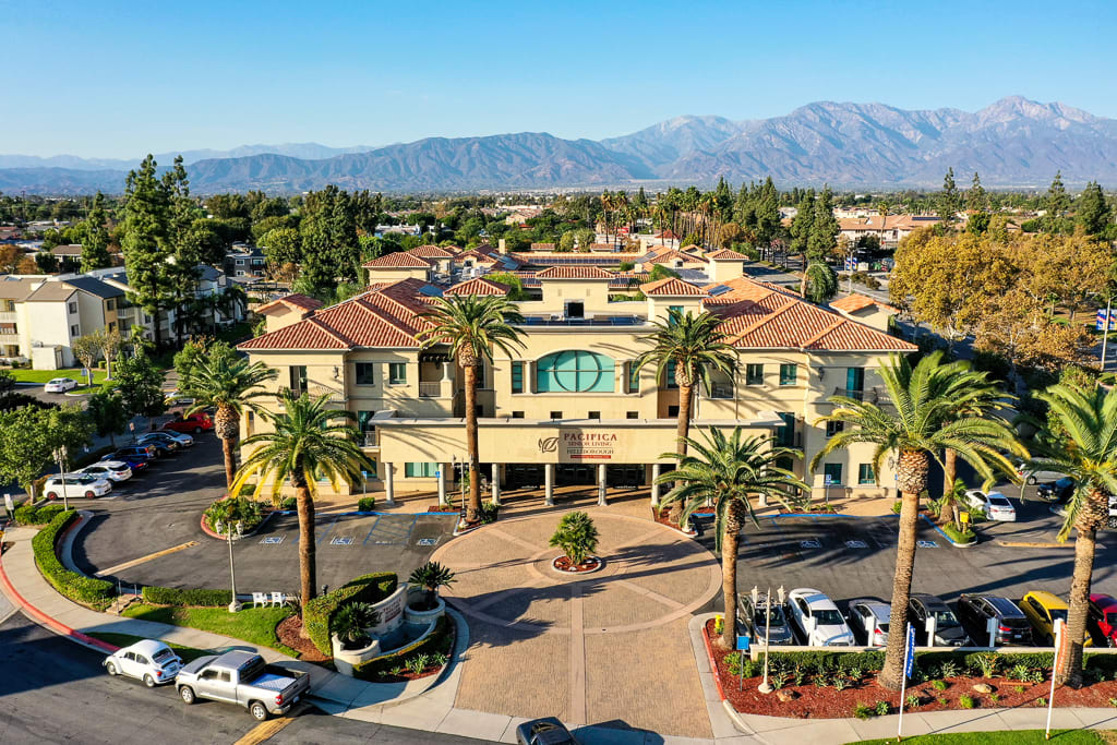 Aerial view of a large building complex surrounded by palm trees with mountains in the background.