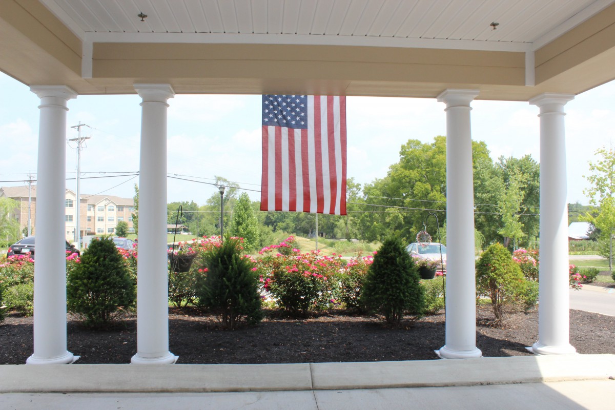 An American flag hanging between two white columns on a patio with greenery in the background.