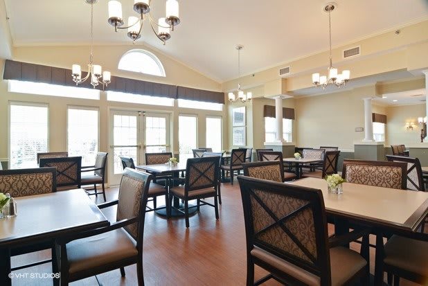 A well-lit dining room with multiple tables and chairs, featuring large windows and elegant chandeliers.