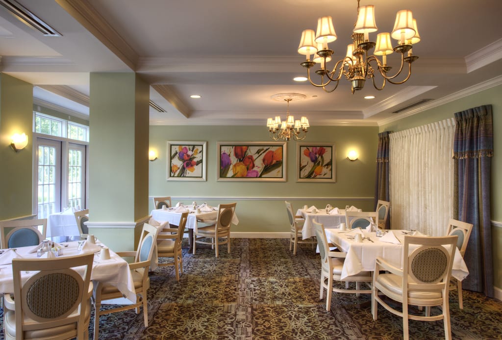 A well-decorated restaurant dining room with neatly arranged tables and chairs, chandeliers, and floral paintings on the walls.