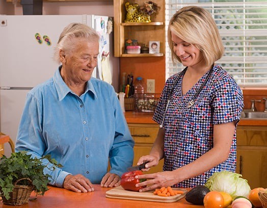 A caregiver assists an elderly person in the kitchen, chopping vegetables together.
