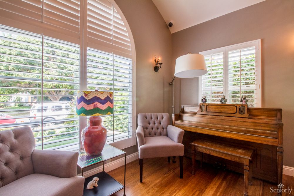 A cozy living room with a piano, armchairs, and a decorative lamp beside large window shutters.