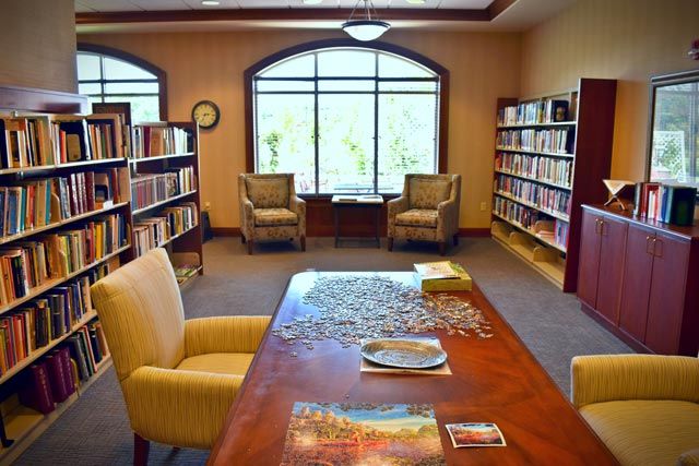 A cozy library room with bookshelves, a table set up for a jigsaw puzzle, and comfortable seating by the window.