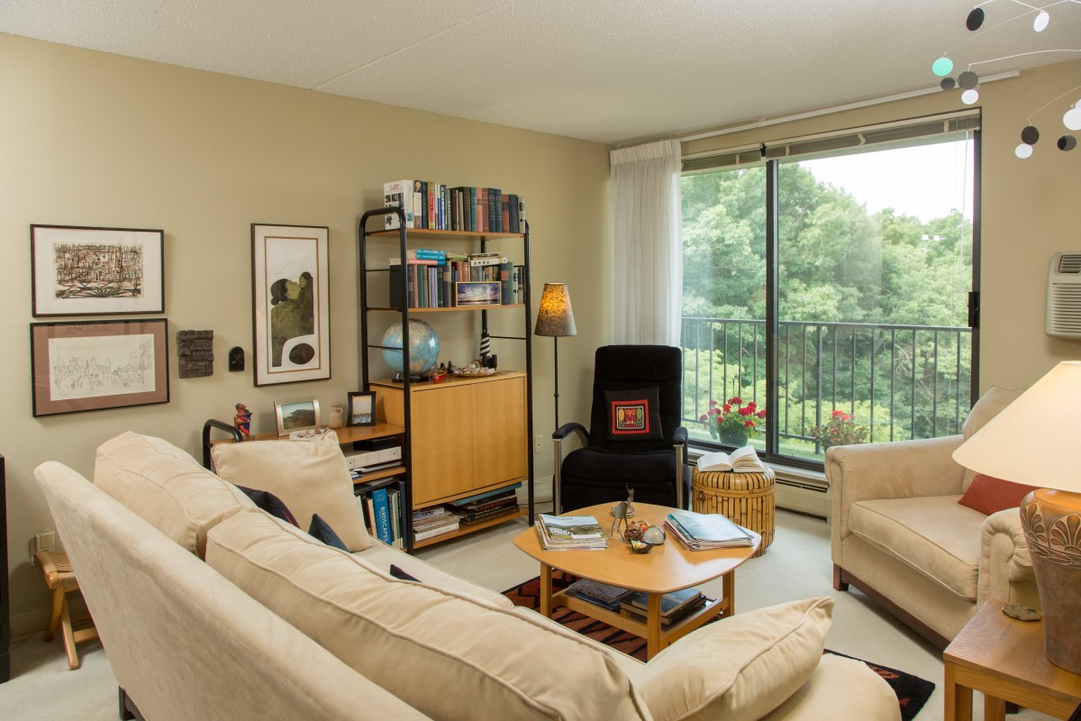 A cozy living room with a comfortable sofa, bookshelf, coffee table, and a large window providing a view of greenery.