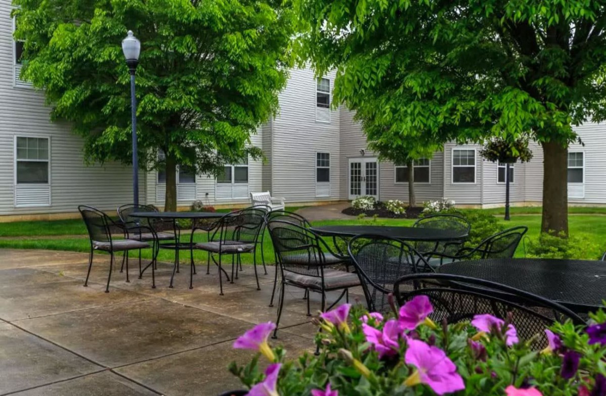 A peaceful outdoor patio area with tables, chairs, and blooming flowers in front of a building surrounded by lush trees.